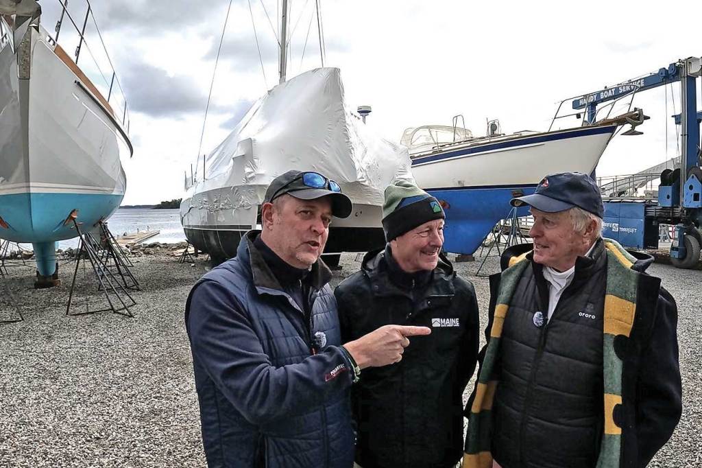 Three men chatting in a boat yard wit boats-on-the-hard in the background.